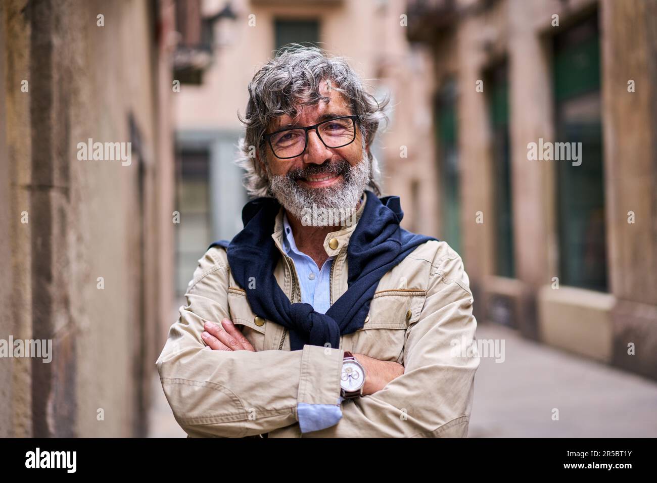 L'uomo anziano dai capelli grigi ha attraversato le braccia sorridendo felice guardando la macchina fotografica in piedi sulla strada della città. Foto Stock