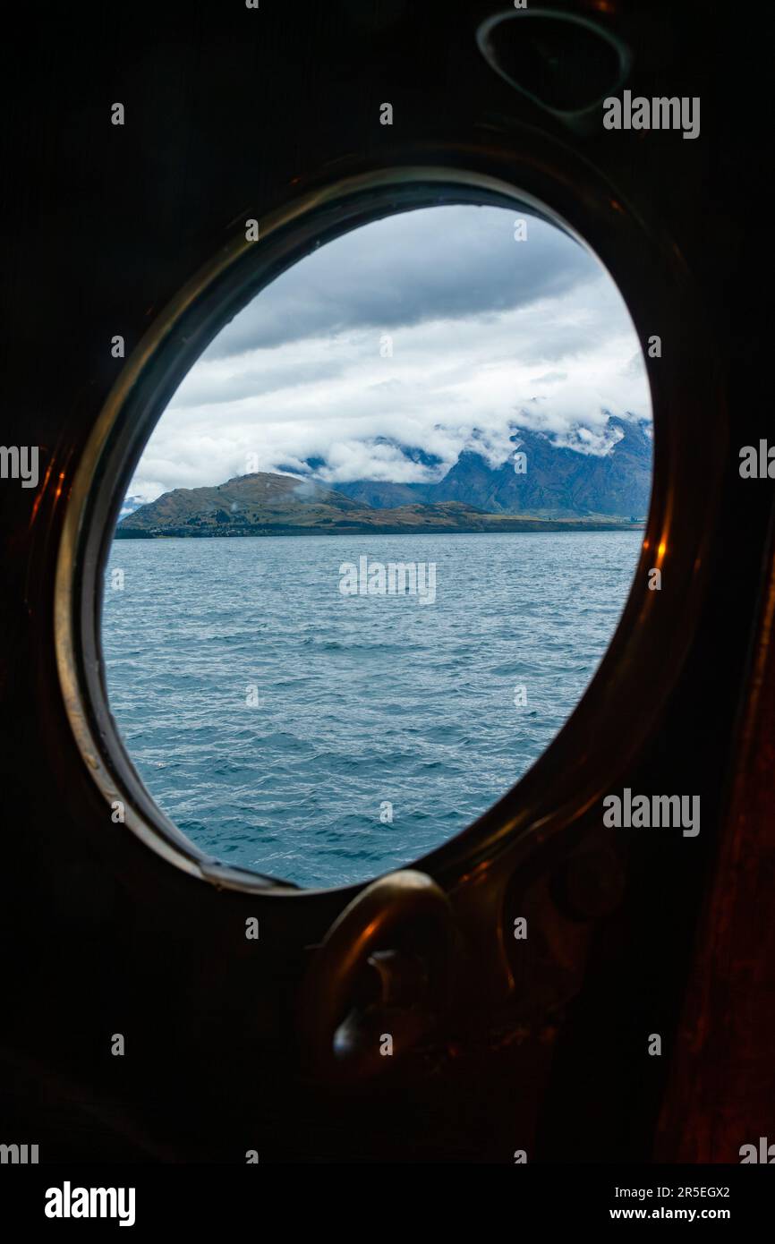 Vista panoramica attraverso il porthole di ottone del battello a vapore turistico che naviga sul Lago Wakatipu nell'Isola Sud della Nuova Zelanda Foto Stock