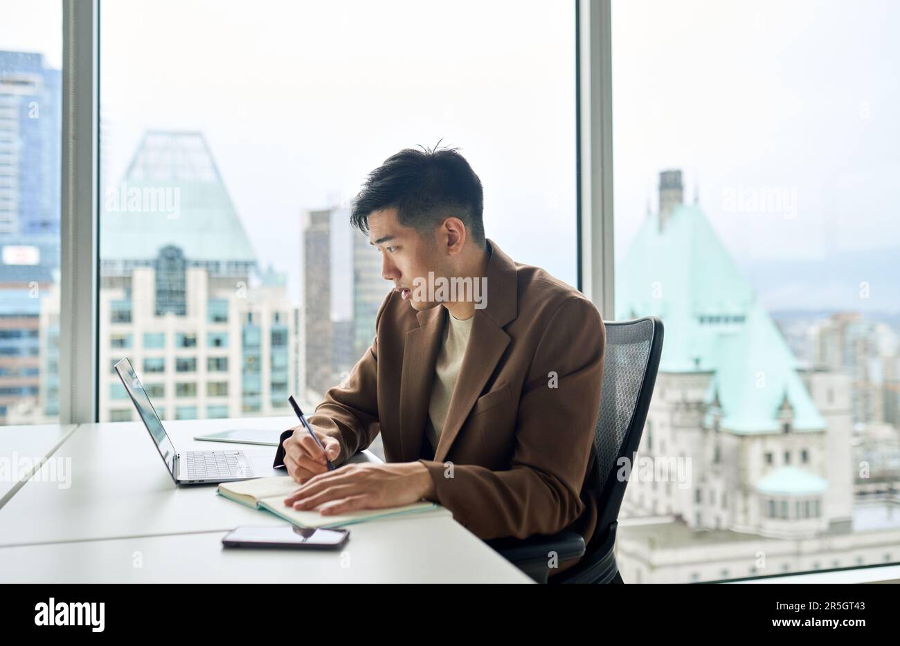 Giovane uomo d'affari asiatico impegnato che lavora su un computer portatile in ufficio scrivendo appunti. Foto Stock