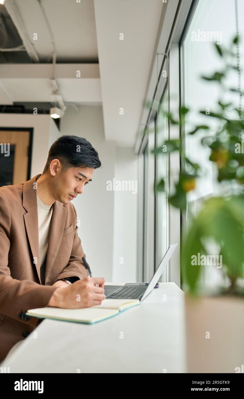 Giovane uomo d'affari asiatico impegnato che lavora su un computer portatile in ufficio scrivendo appunti. Foto Stock