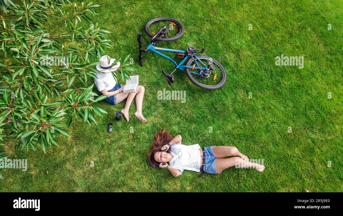 Ragazze giovani con la bicicletta nel parco, utilizzando computer portatile e cuffie, due ragazze studentesche che studiano online all'aperto seduti sull'erba vicino alla bicicletta Foto Stock