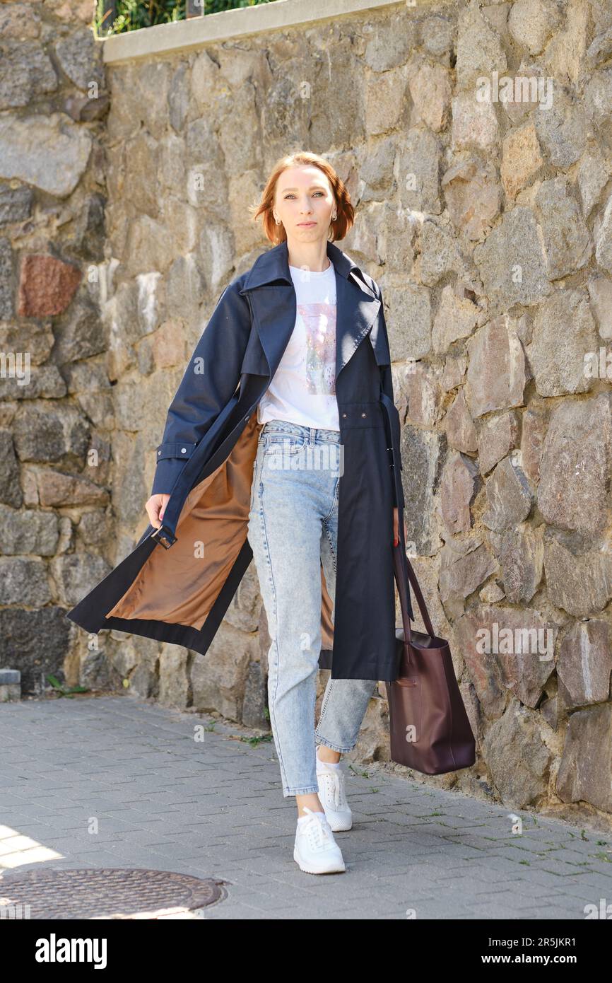 Elegante donna di mezza età cammina per la strada della città indossando un trench blu, jeans e sneakers Foto Stock