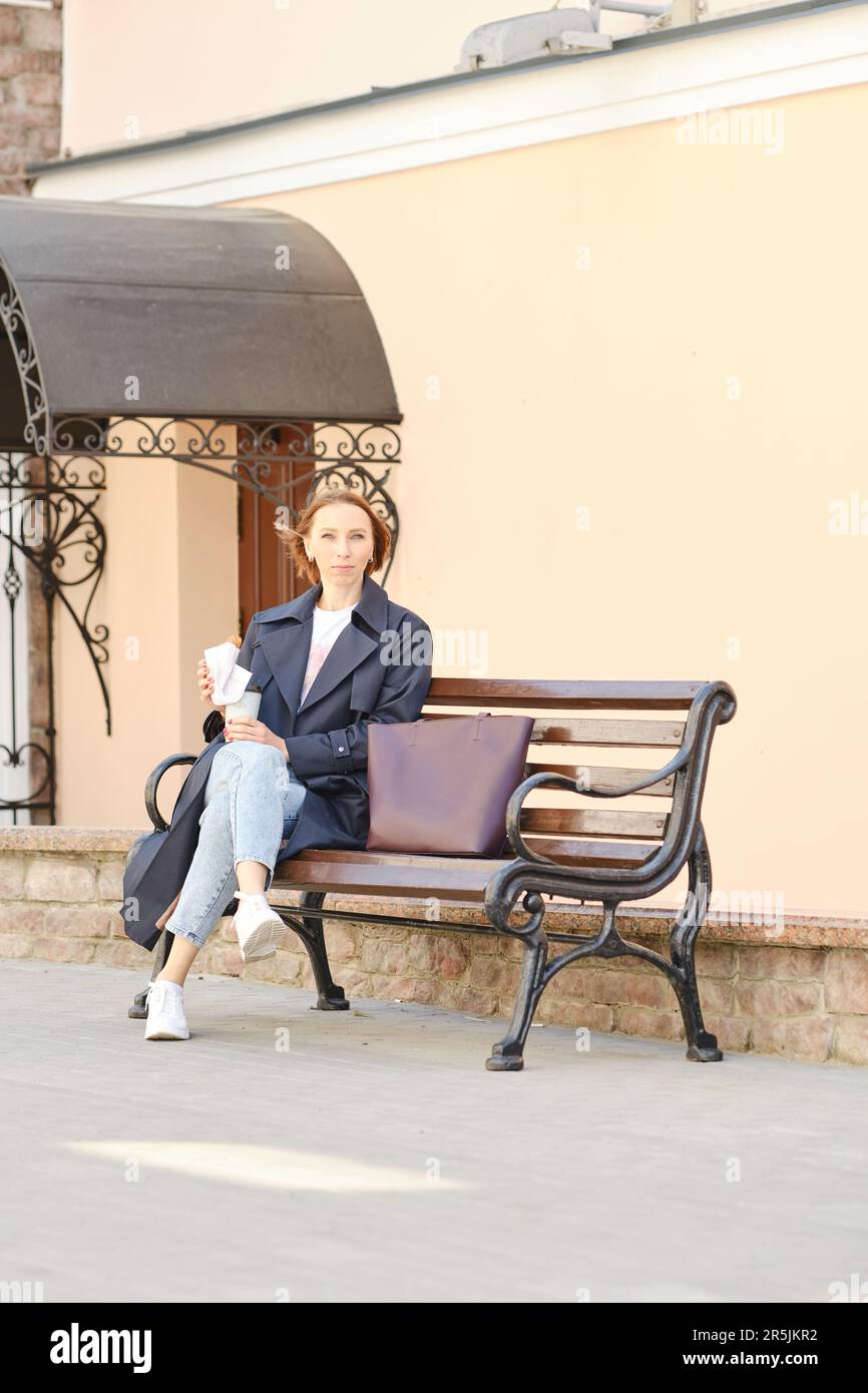 Elegante donna d'affari con testa rossa in trench coat e tazza da caffè seduta sulla panca Foto Stock