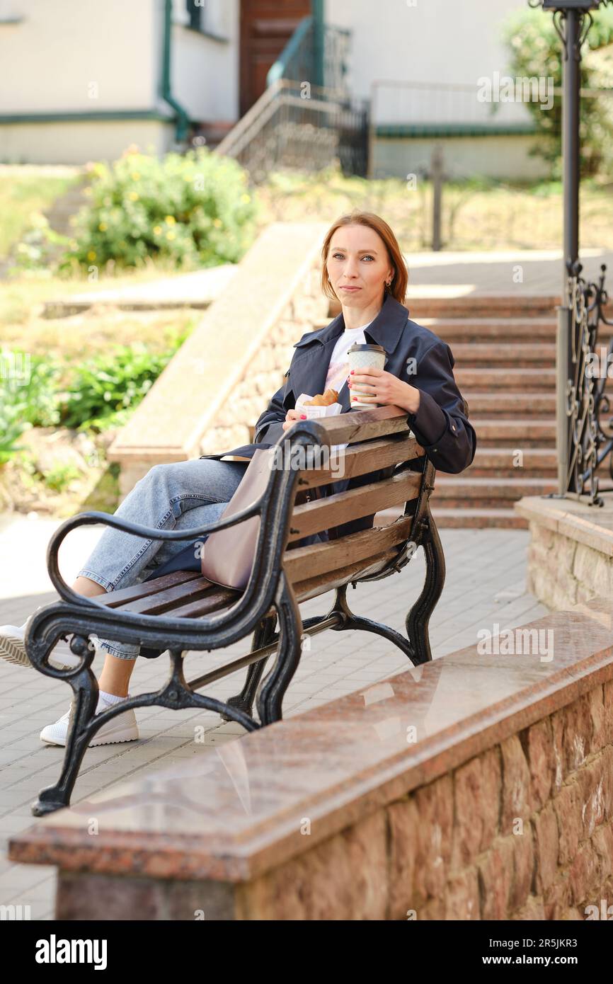 Donna di mezza età dai capelli rossi seduta sulla panchina che tiene il caffè e croissant che si diverte Foto Stock