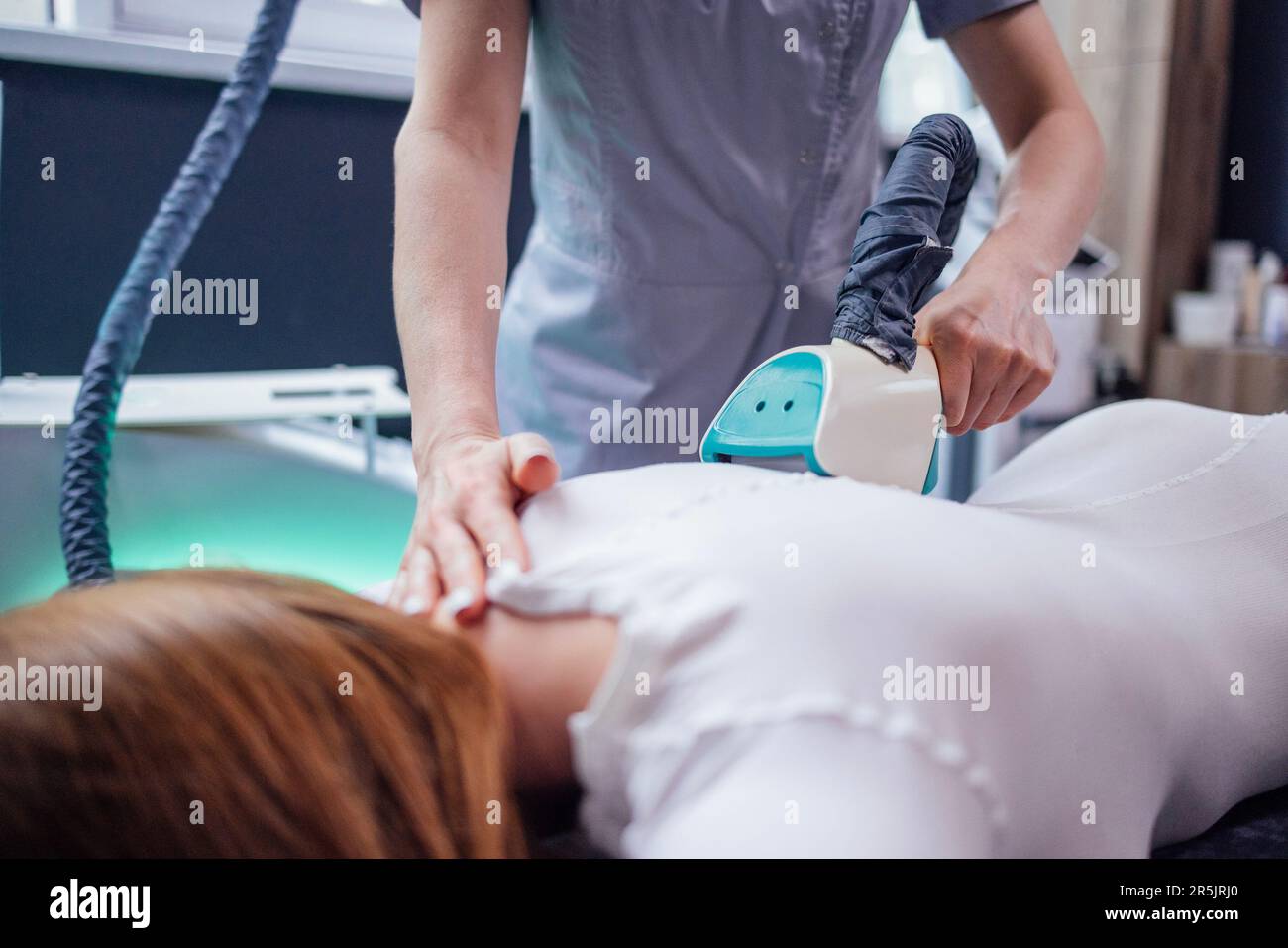 Cosmetician che usa l'apparecchio di dimagrimento del GPL sul corpo della cliente femminile in tuta bianca che giace sul lettino Foto Stock