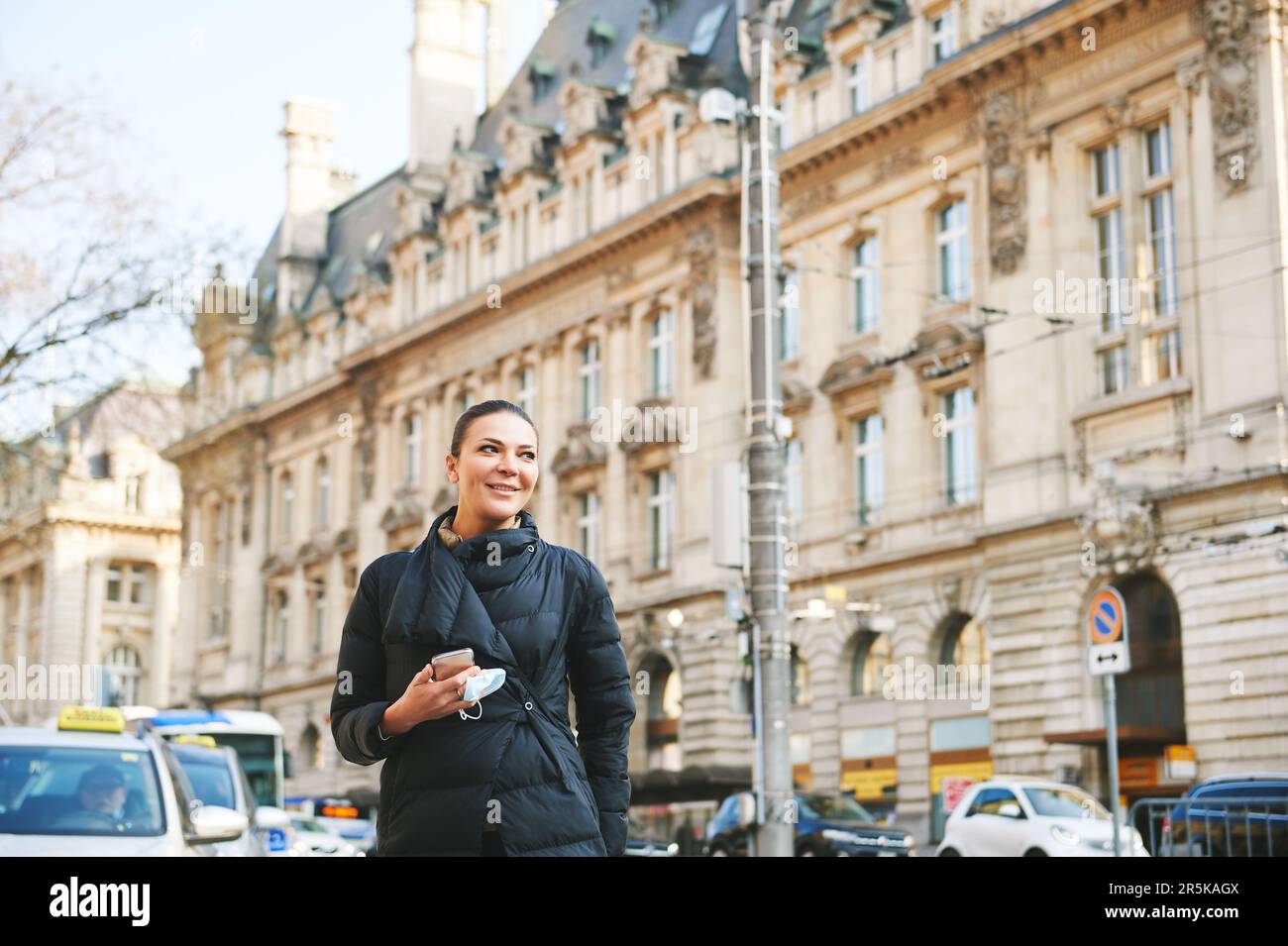 Felice giovane donna che cammina per la strada, indossa una giacca nera calda, tiene in mano il telefono e la maschera facciale Foto Stock