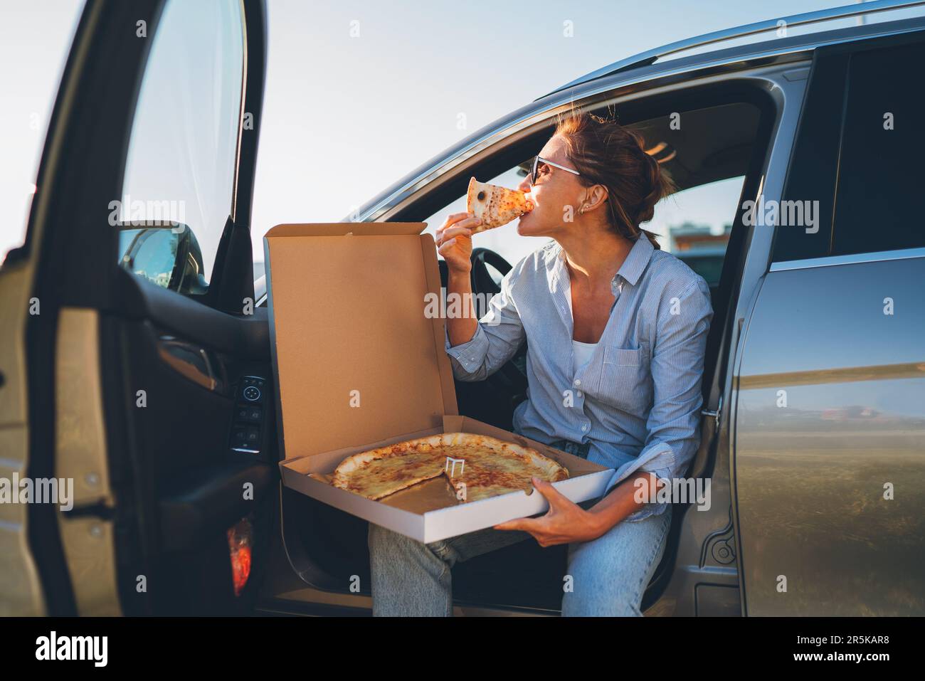 Donna anziana che mangia pizza italiana appena cotta seduto sul sedile dell'autista durante la pausa pranzo e godendosi la luce del tramonto. Auto in viaggio, fast food ea Foto Stock