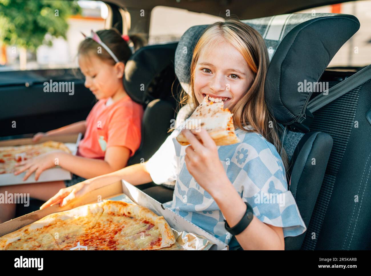 Ritratto di una ragazza sorridente positiva che mangia una pizza italiana appena cotta seduta con una sorella sul sedile posteriore dell'automobile nei seggiolini dell'automobile del bambino. Buona infanzia, fastfoo Foto Stock