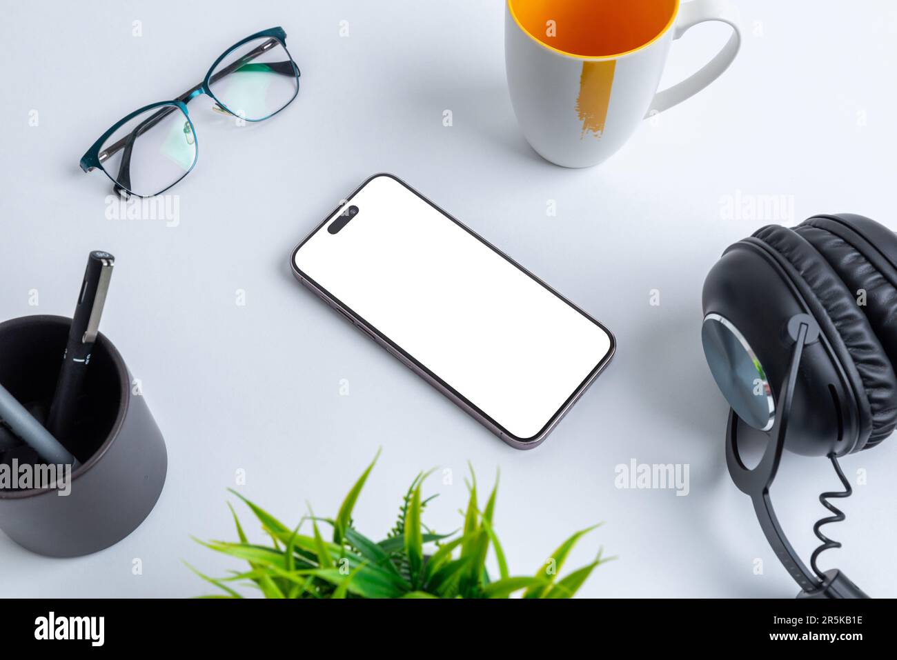 Mockup telefonico con schermo isolato per la presentazione delle applicazioni collocato sulla scrivania dell'ufficio, circondato da tazza da caffè, bicchieri, penne, piante e cuffie. Foto Stock