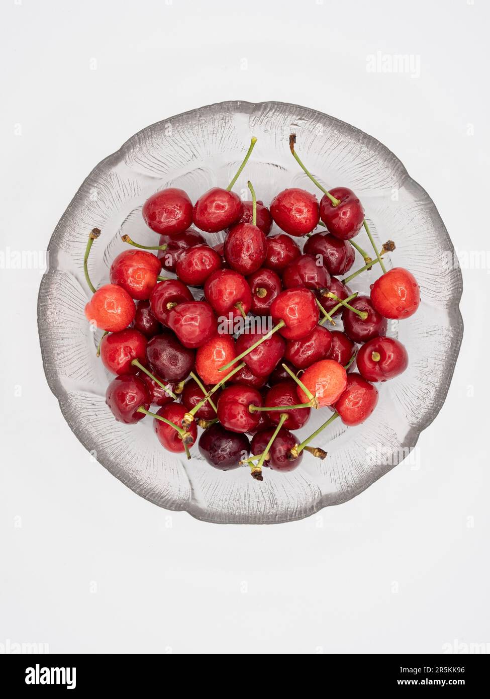 Ciliegie rosse organiche, scintillanti con gocce d'acqua rinfrescanti, presentate elegantemente in un recipiente di vetro incontaminato, perfettamente isolate su un bac bianco pulito Foto Stock