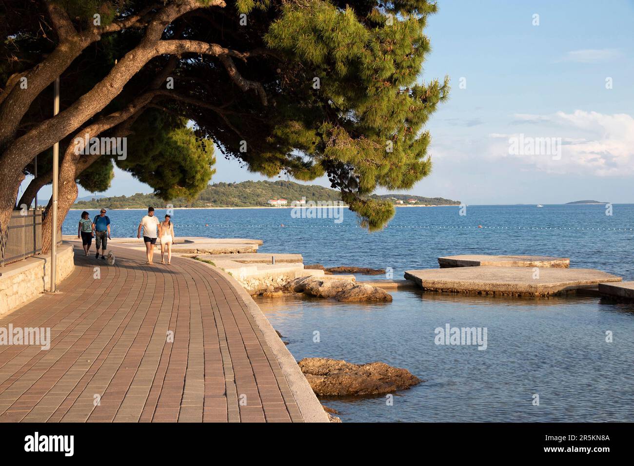 Vodice, Croazia - 30 maggio 2023: Passeggiata con pini sul mare e la gente che cammina in una giornata di sole al tramonto Foto Stock