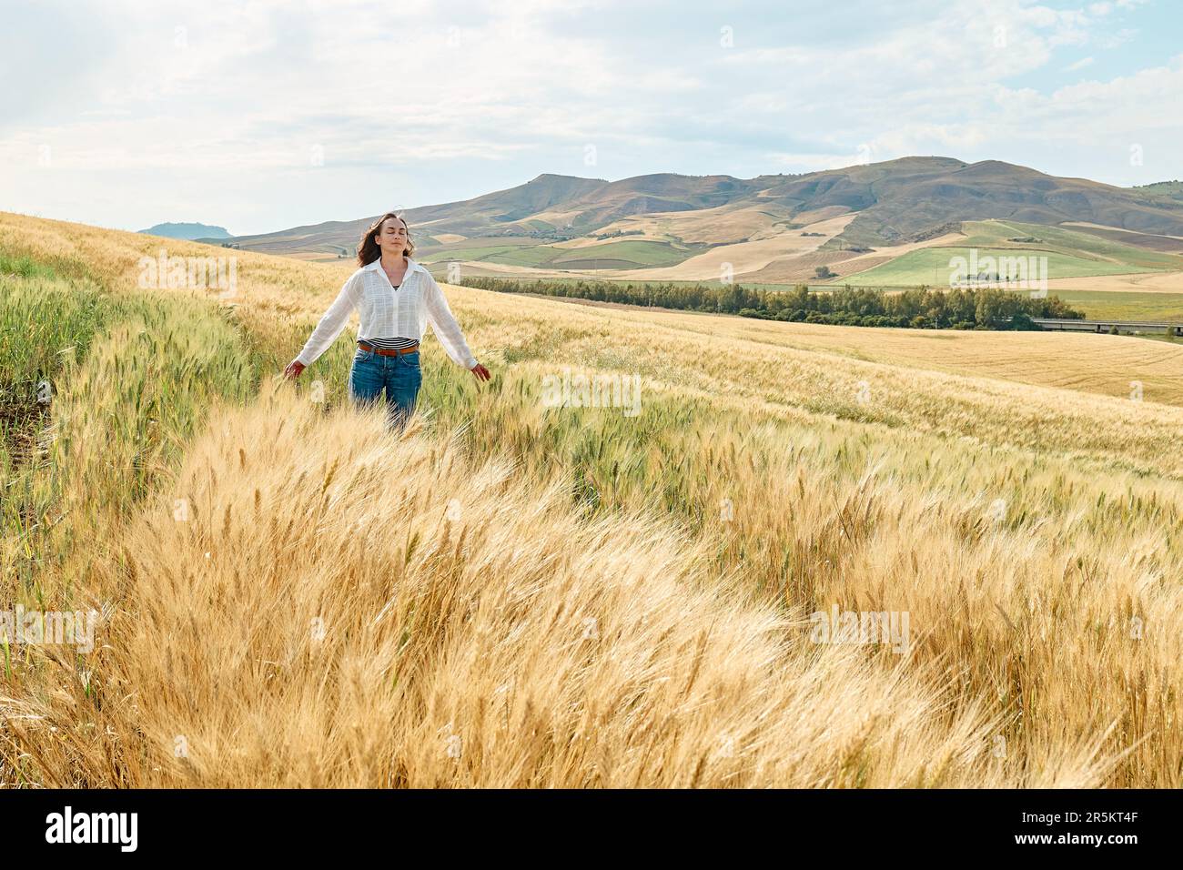 Donna che cammina in campo di grano dorato in caldo sole estivo e cielo blu con nuvole bianche con montagne paesaggio collinare sullo sfondo. Foto Stock