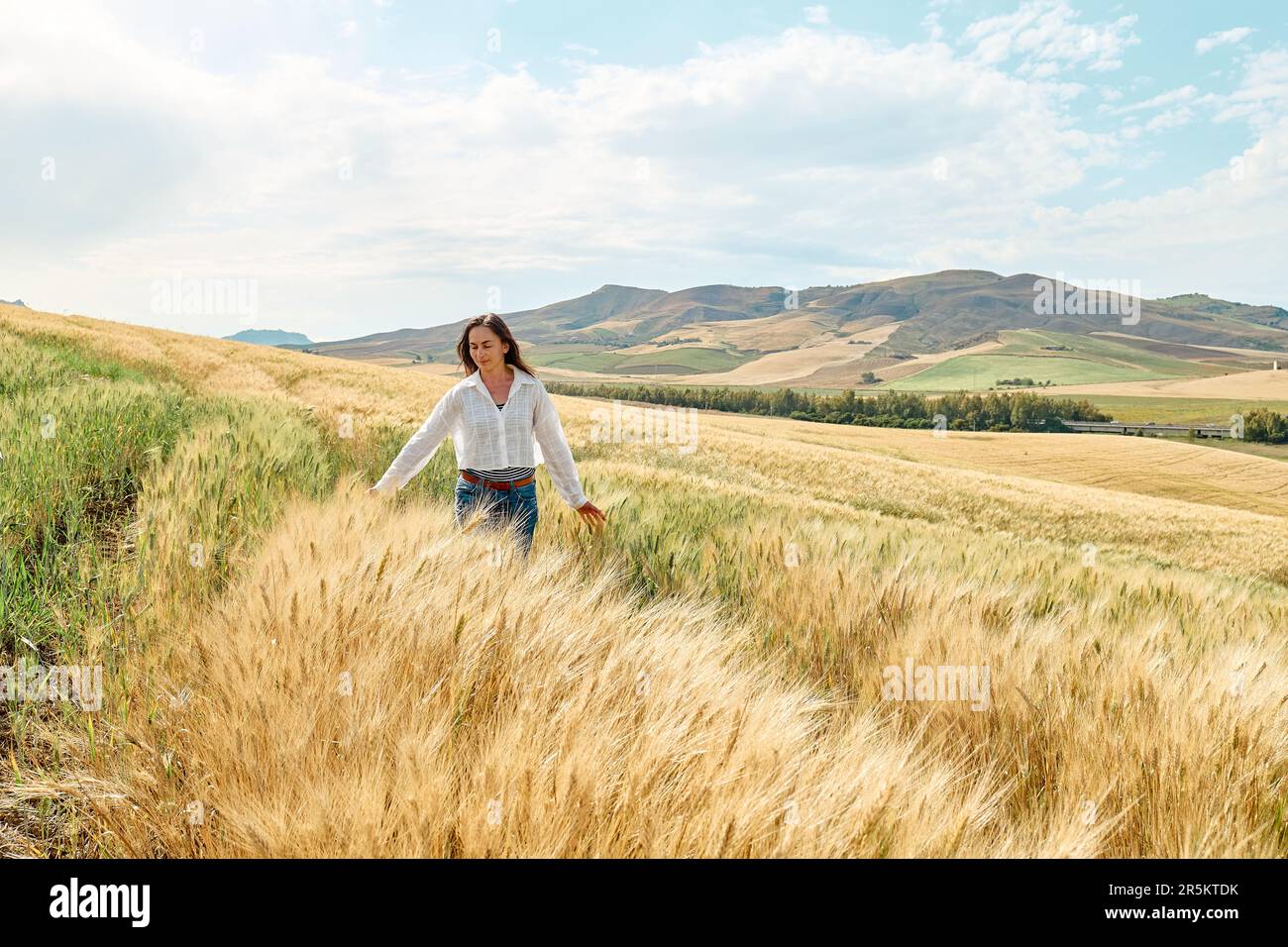 Donna che cammina in campo di grano dorato in caldo sole estivo e cielo blu con nuvole bianche con montagne paesaggio collinare sullo sfondo. Foto Stock