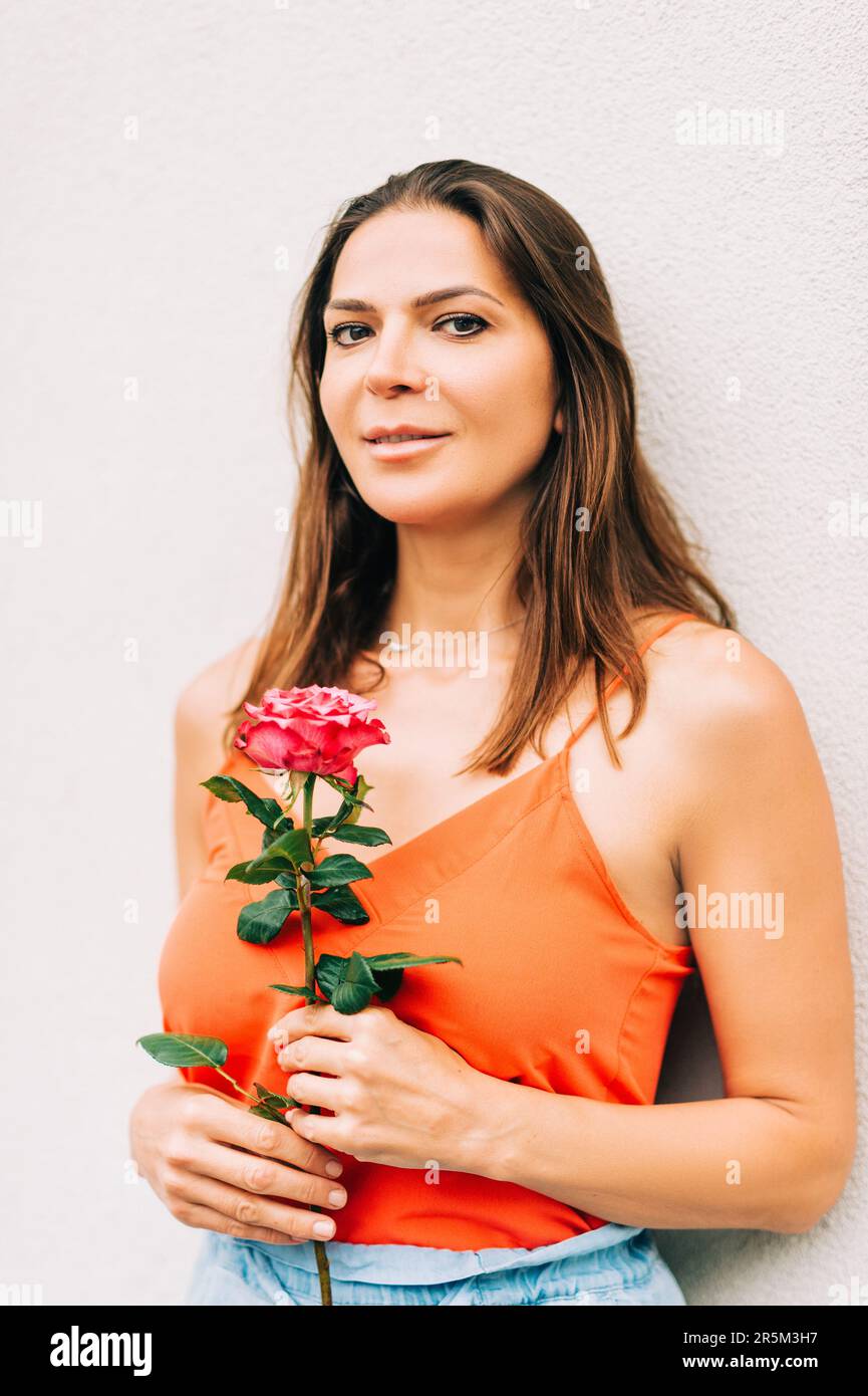 Ritratto all'aperto di attraente donna che tiene fiore rosa, indossando abiti arancioni Foto Stock