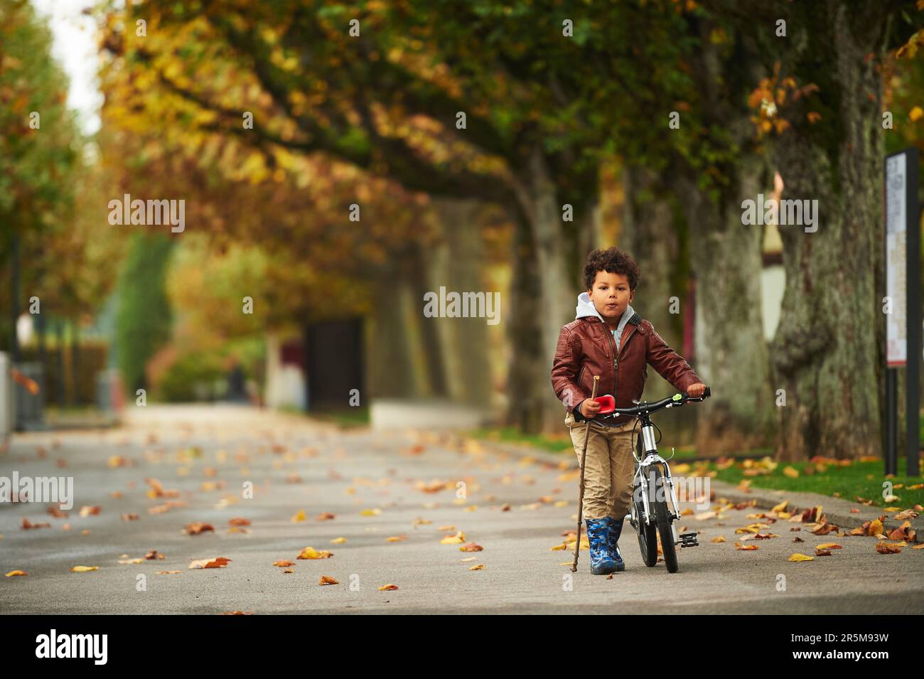 Carino ragazzino che cammina nel parco autunnale, tira la moto, tiene un bastone Foto Stock