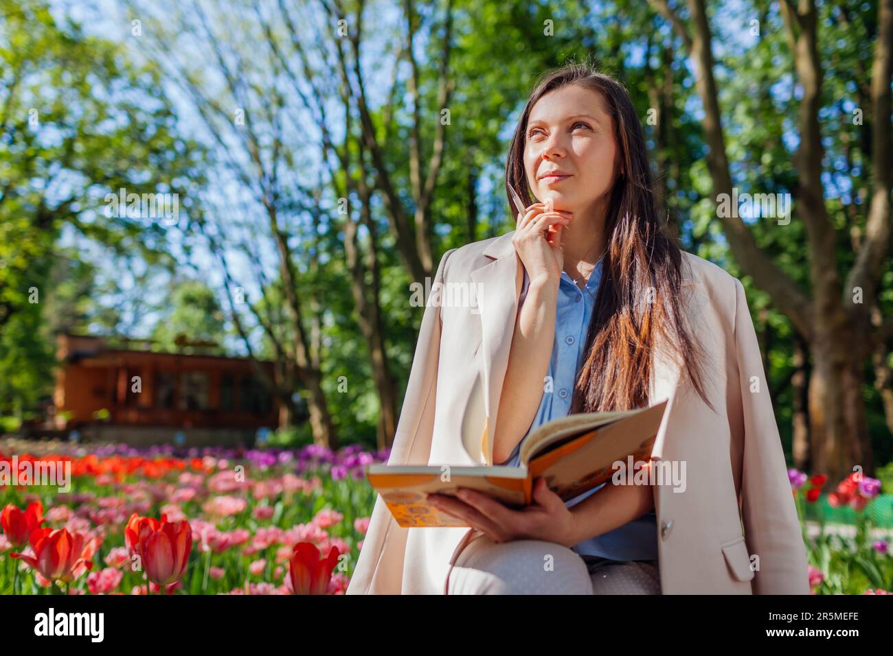 Giovane paesaggista caucasico pensare tenendo notebook in primavera parco tra fiori fiorenti. Donna che scrive le idee. Spazio. Spazio Foto Stock