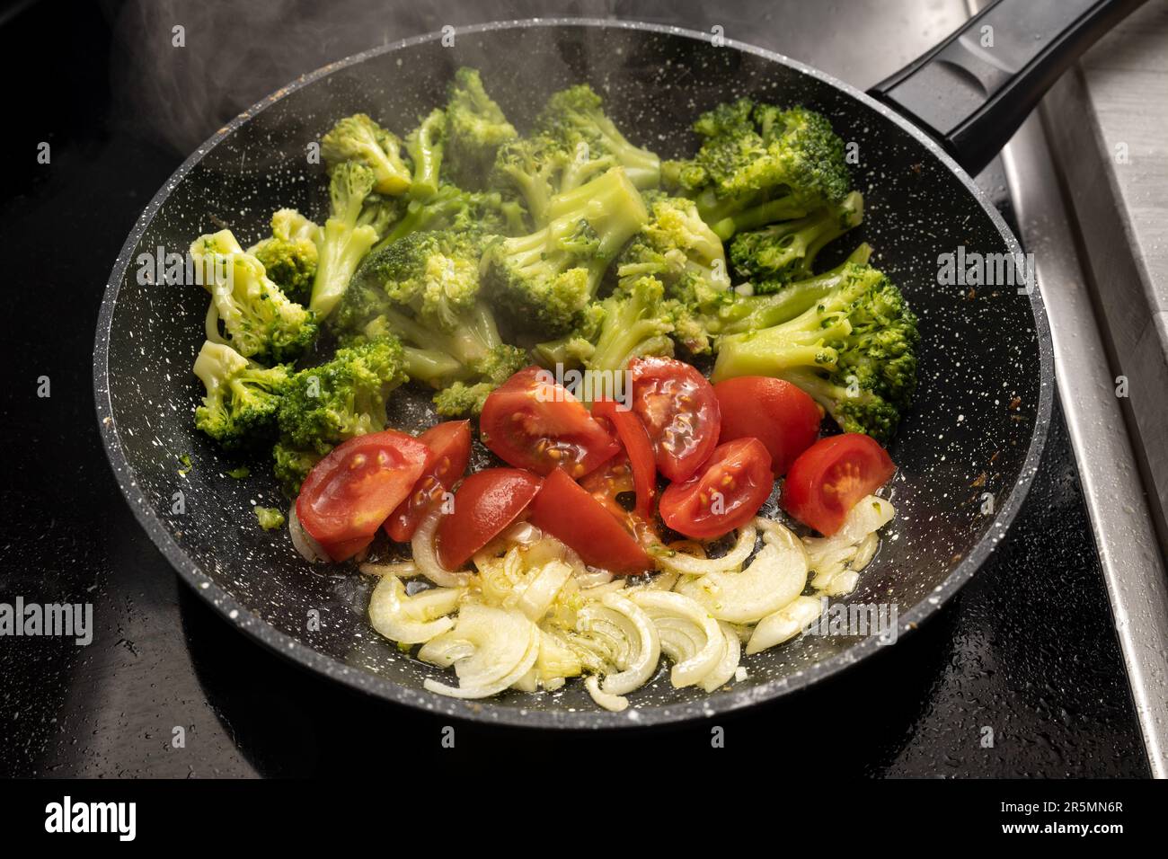 Cipolle, pomodori e broccoli vengono saltati in una padella fumante sul piano cottura nero per un pasto vegetariano sano, focus selezionato, profondità ridotta o Foto Stock