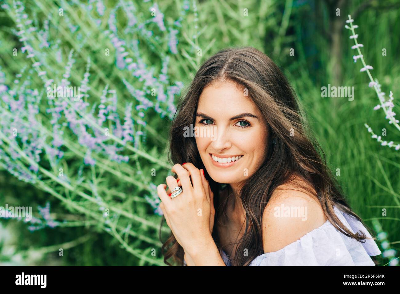 Primo piano esterno ritratto di bella giovane donna con capelli scuri in posa giardino estivo con fiori viola Foto Stock