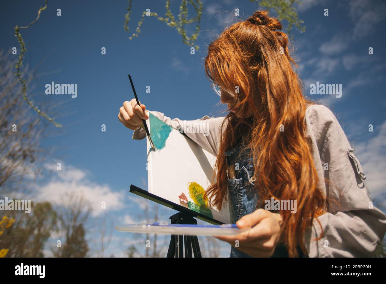 La ragazza dello zenzero sta dipingendo su un cavalletto con un pennello in mano mentre sta in piedi al parco Foto Stock