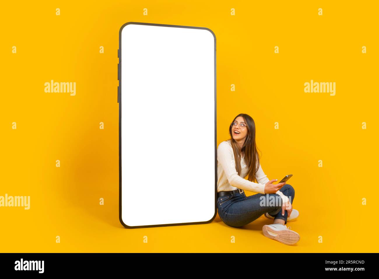 Mockup per la progettazione di applicazioni. Ragazza caucasica seduta vicino a un grande cellulare con schermo vuoto. Utilizzando il telefono cellulare. Visualizzazione enorme. Foto Stock