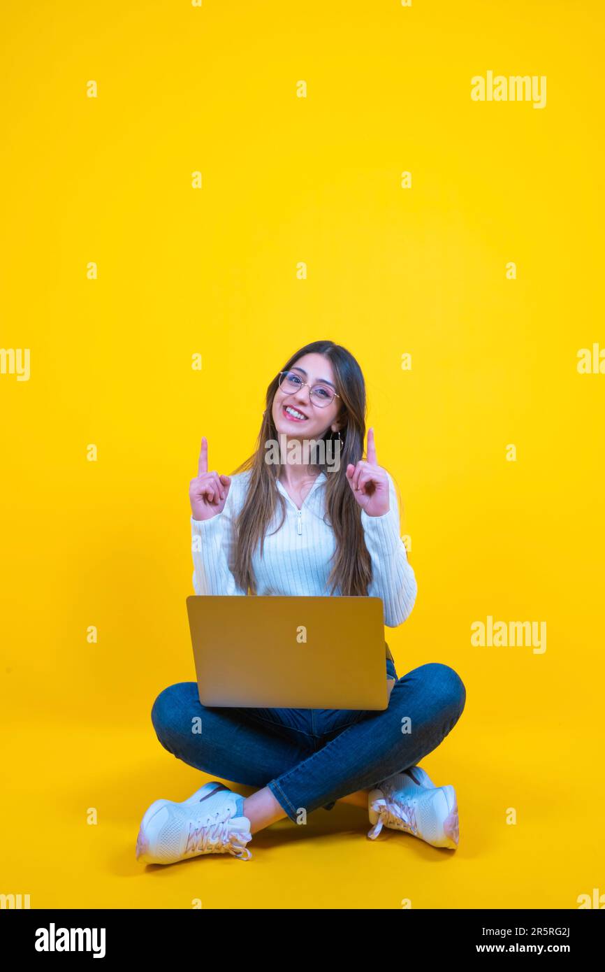 Guarda in alto, donna felice ed eccitata seduta a terra con un notebook e punta verso l'alto. Visualizzazione dello spazio di copia. Studio isolato sfondo arancione. Foto Stock