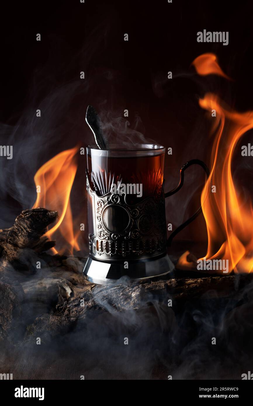 Un bicchiere di tè nel portabicchieri e lingue da gioco e riflessi di fiamma con fumo sullo sfondo su legno driftwood Foto Stock