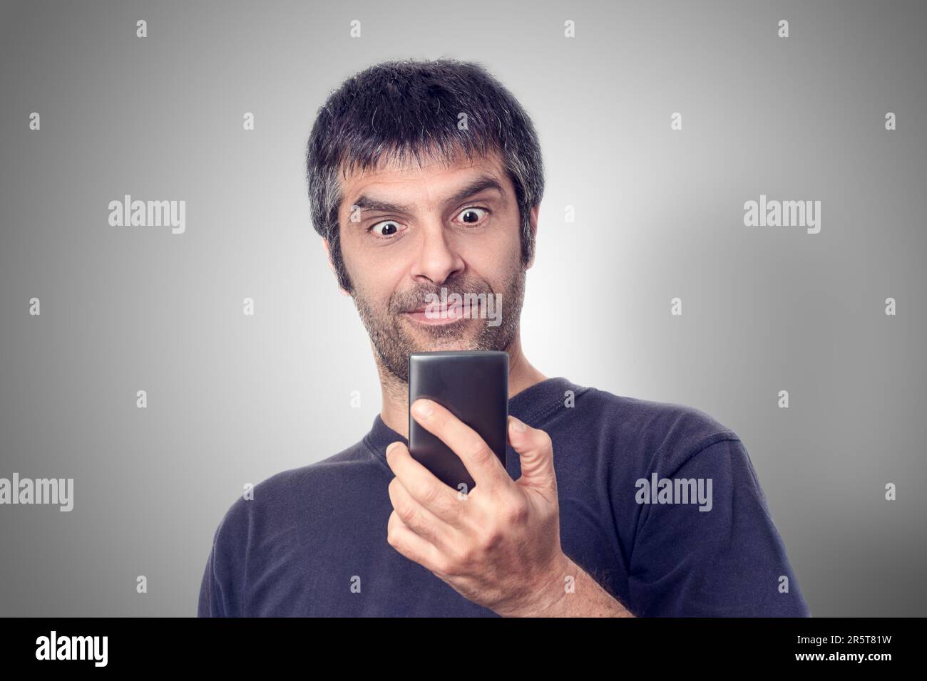 Uomo maturo con capelli scuri che indossa un'espressione sorpresa mentre guarda il suo telefono cellulare su sfondo grigio. Foto Stock