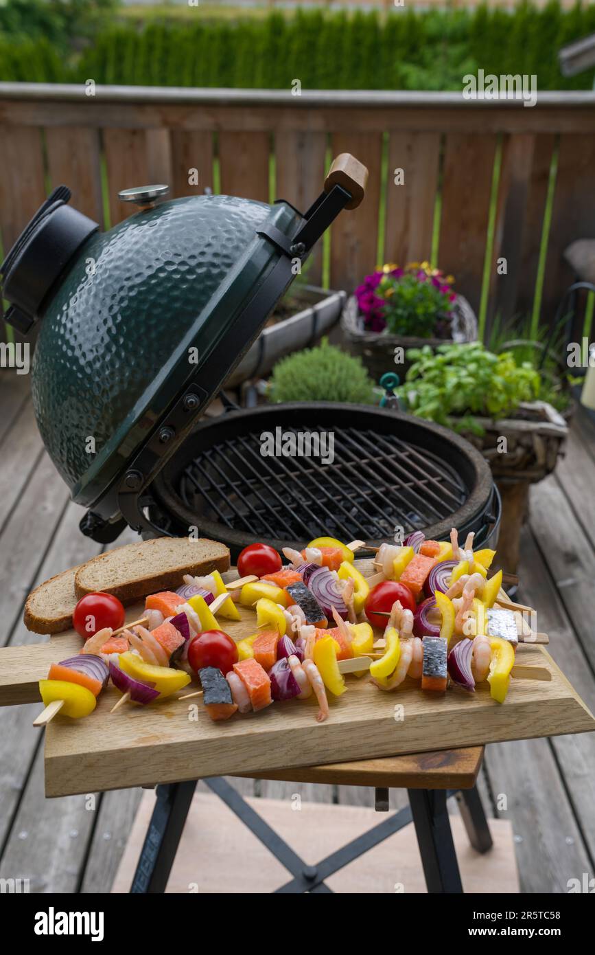 spiedini di gamberetti crudi con salmone e verdure su un asse di legno per grigliare su una griglia a carbone sul balcone in una giornata estiva Foto Stock