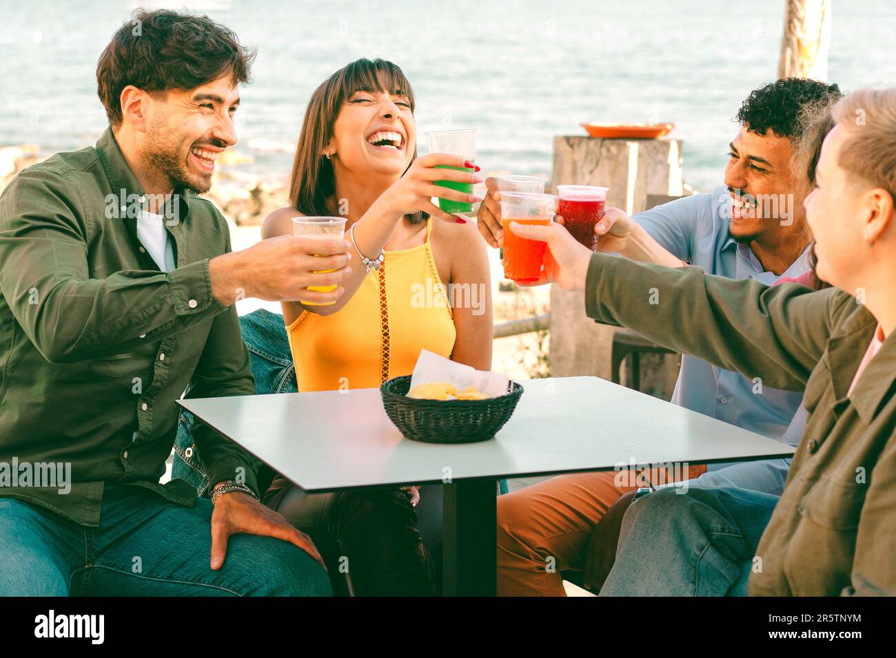 Un gruppo diversificato di amici seduti in un bar sulla spiaggia, brinda allegramente con vibranti cocktail in tazze di plastica. La vivace scena mostra la gioia Foto Stock