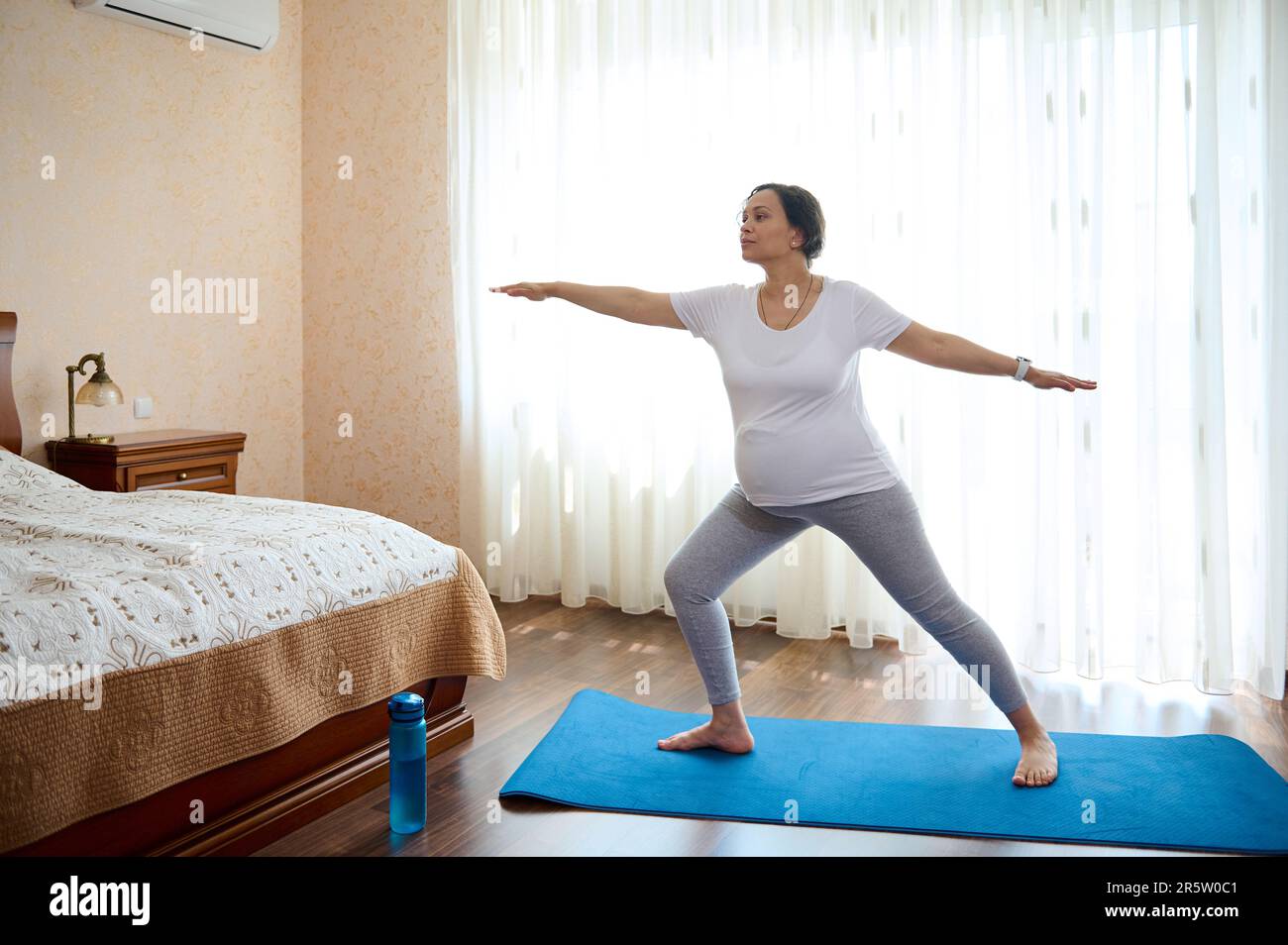 Donna incinta in gravidanza tardiva, pratica yoga a casa, in piedi in guerriero posa sul materassino di esercizio. Verticale a lunghezza intera Foto Stock