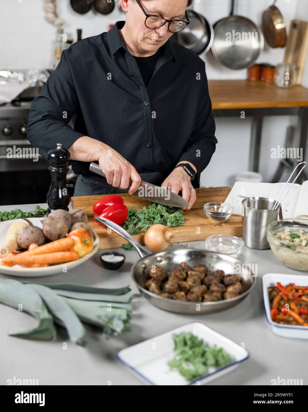Un uomo anziano sta preparando un pasto ad un tavolo da cucina, usando una varietà di attrezzi di taglio Foto Stock