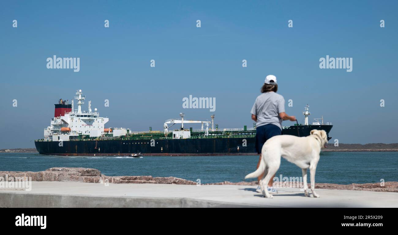 La nave petrolifera naviga sull'acqua blu di un canale di spedizione e passa oltre il marciapiede, il molo di persona, il molo di cani e rocce. Foto Stock