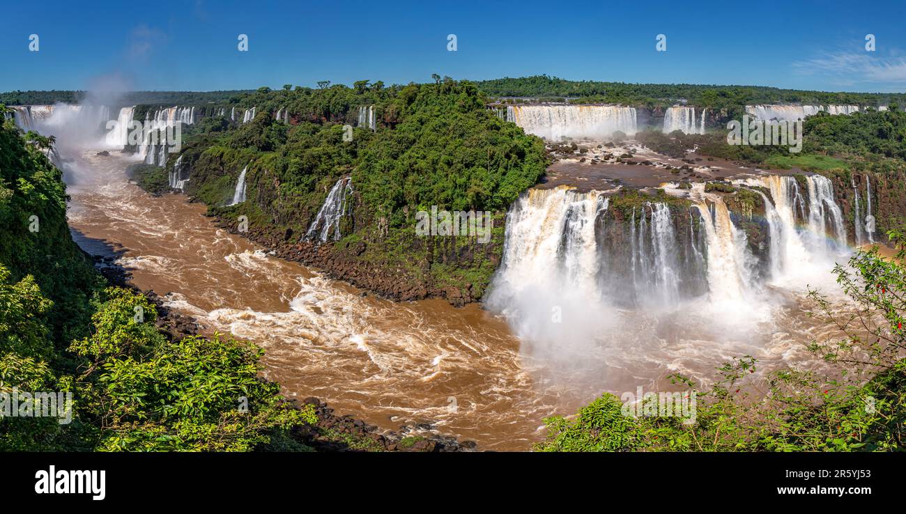 Vista panoramica della gola delle cascate di Iguazu con il fiume marrone, le cascate bianche e la lussureggiante foresta pluviale al sole Foto Stock