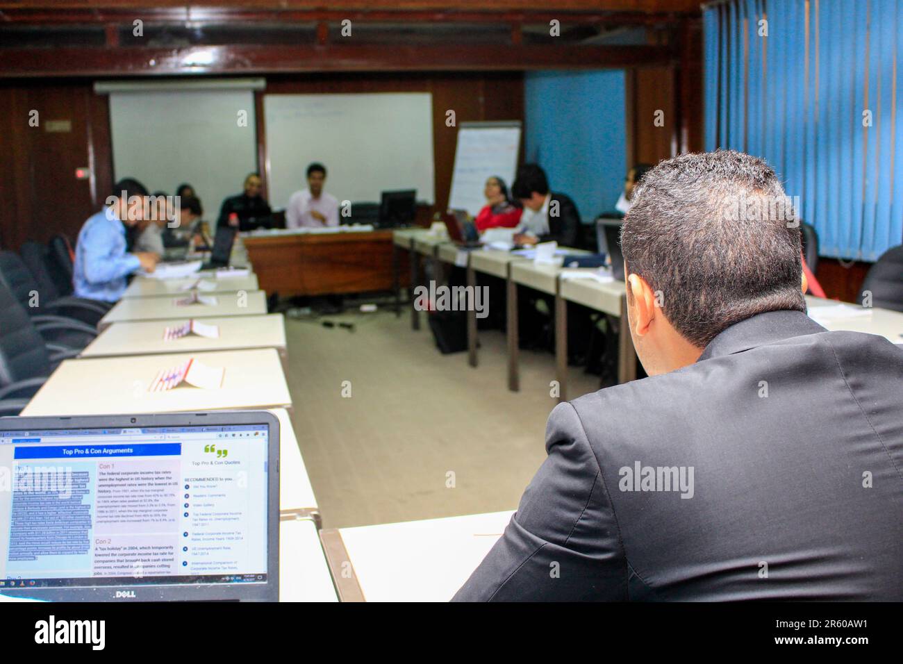 Riunione di lavoro professionale in una sala conferenze. Le persone sono sedute intorno a un tavolo a U con i laptop aperti e impegnati in discussioni. Un notebook sc Foto Stock