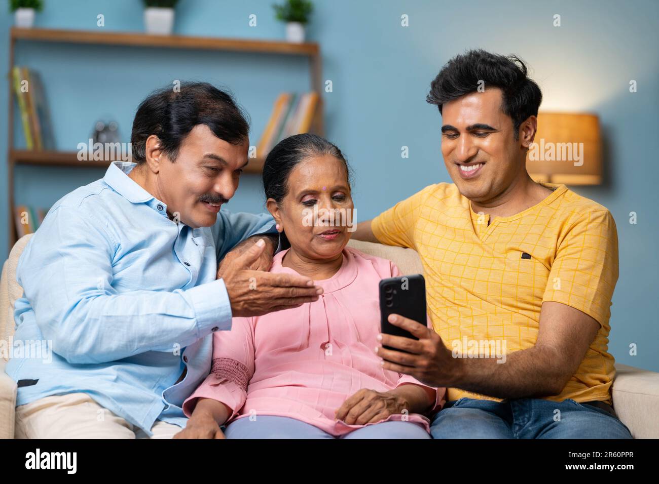 Figlio adulto indiano felice che mostra il telefono cellulare ai genitori anziani mentre si siede sul divano a casa - concetto di applicazione matrimonio, tecnologia e legame familiare Foto Stock