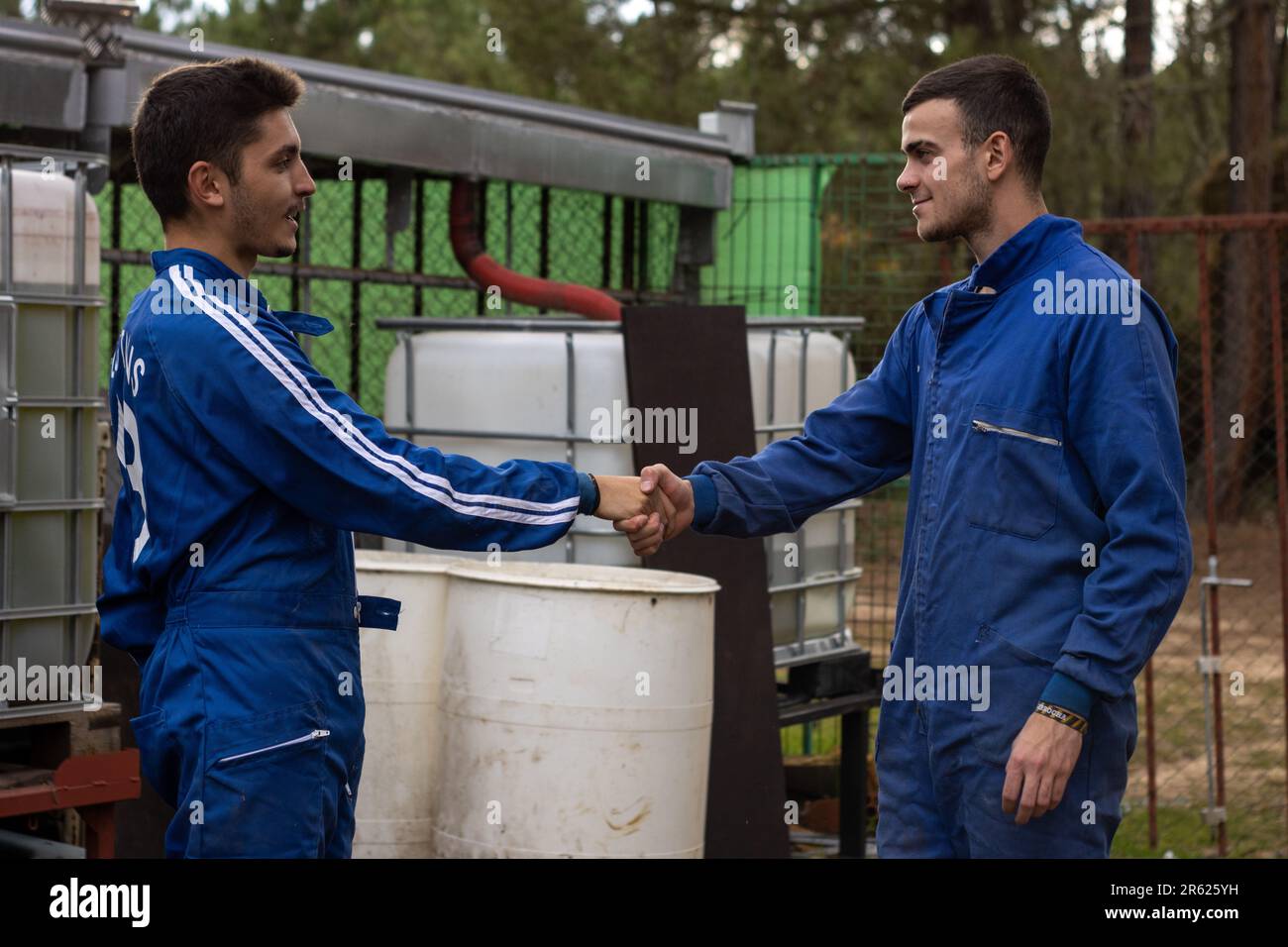 Due uomini caucasici nei loro anni venti indossando abbigliamento casual business scuotendo le mani in una zona boscosa con un rustico barile di legno e recinzione di metallo nella b Foto Stock