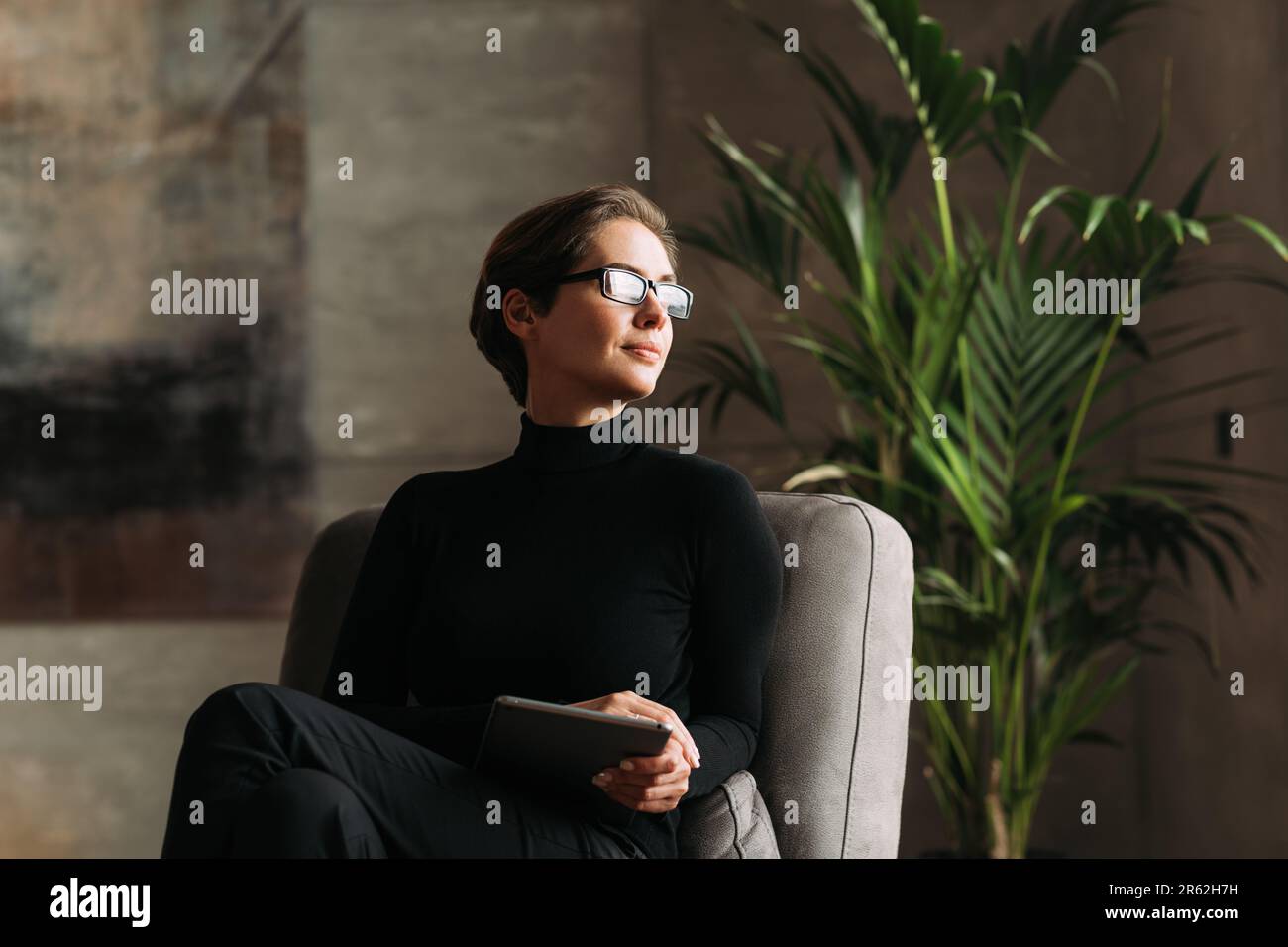 Giovane donna in nero abbigliamento formale indossando occhiali che tengono un tablet digitale mentre si siede nel suo loft Foto Stock