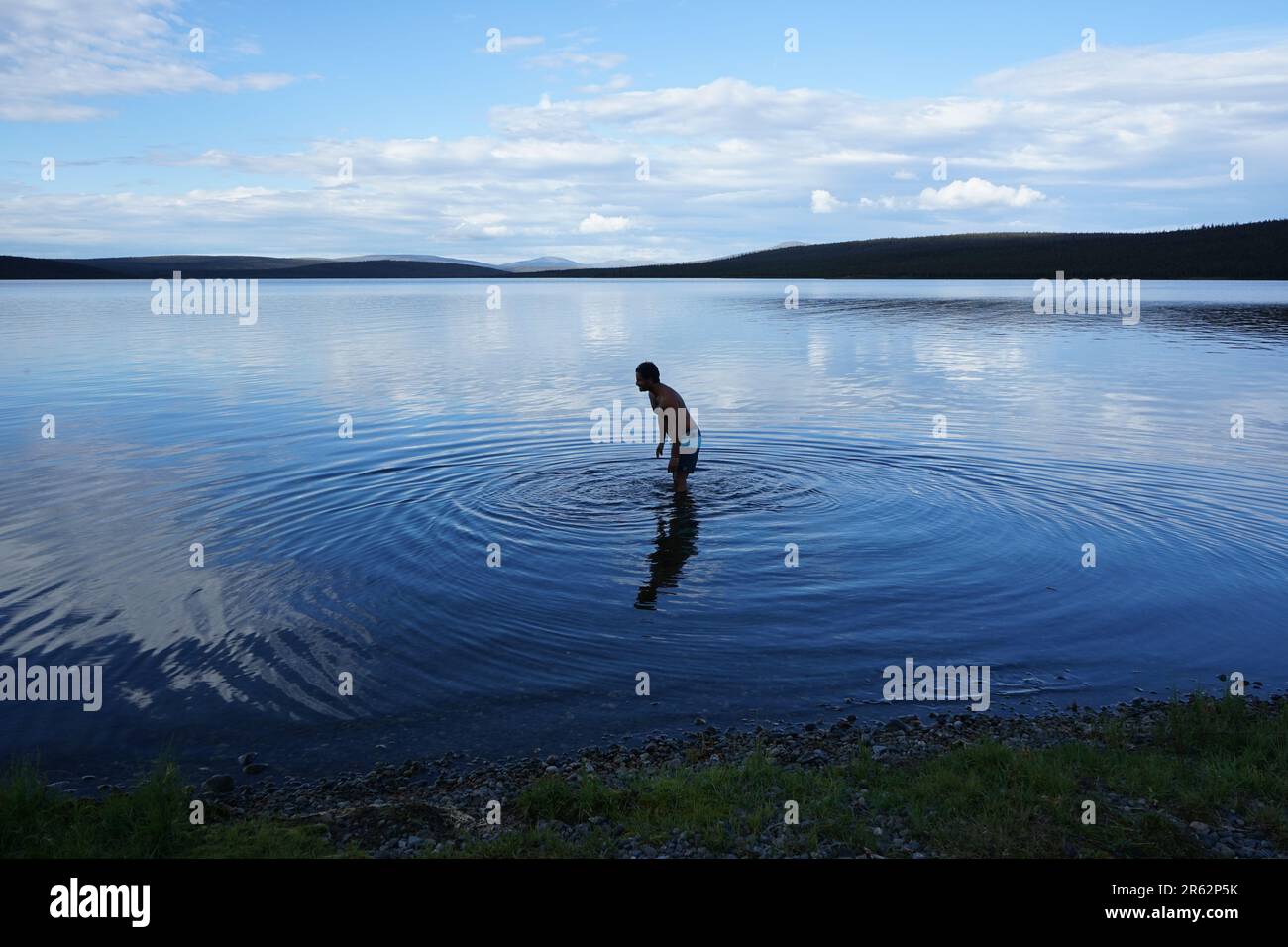 Una serena vista sul lago con acque cristalline e fredde. Un uomo si erge sul lago, creando ondulazioni circolari, abbracciando la bellezza dei dintorni remoti. Foto Stock