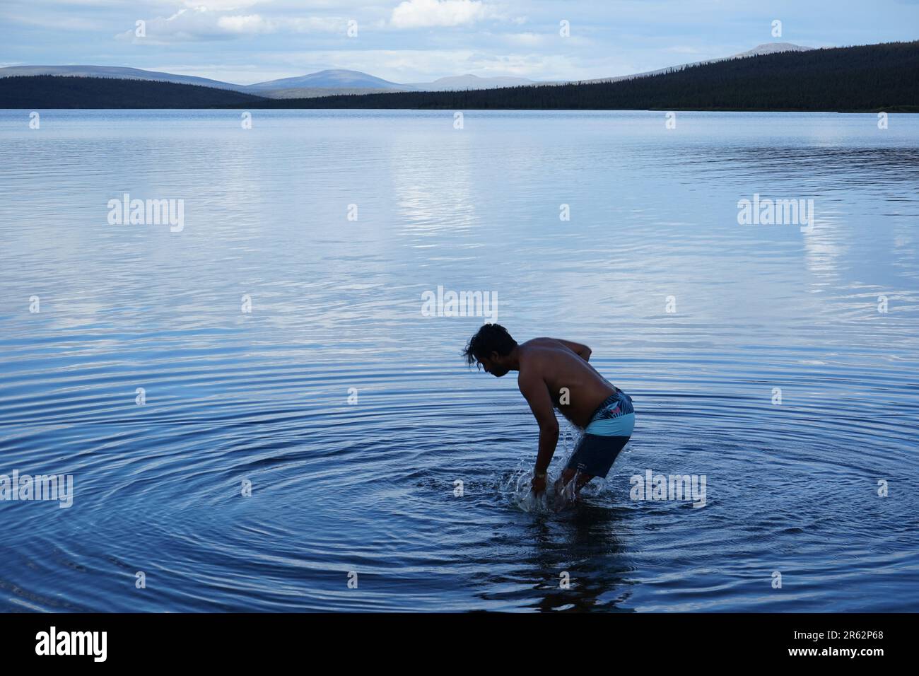Una serena vista sul lago con acque cristalline e fredde. Un uomo si erge sul lago, creando ondulazioni circolari, abbracciando la bellezza dei dintorni remoti. Foto Stock