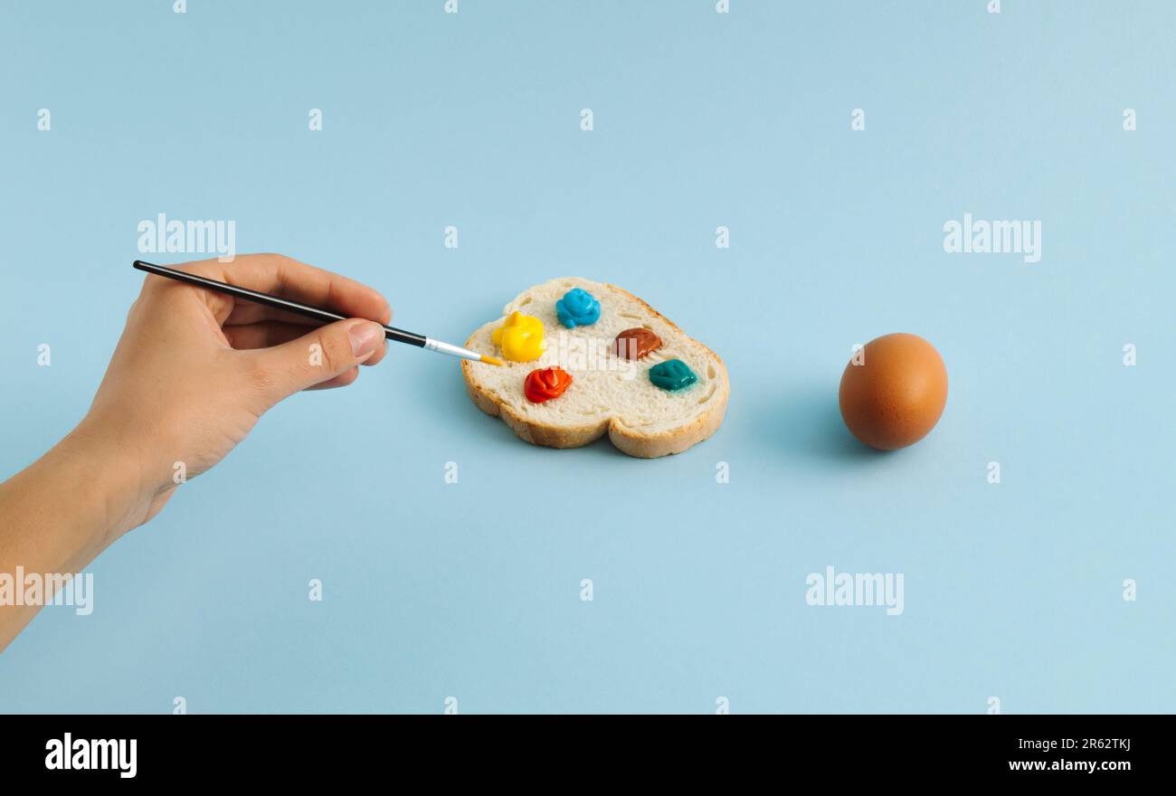 Un uovo bollito in mano e una fetta di pane con tavolozza colorata su sfondo blu pastello. La donna sta colorando le uova per la festa di Pasqua. Pasqua creativa c Foto Stock