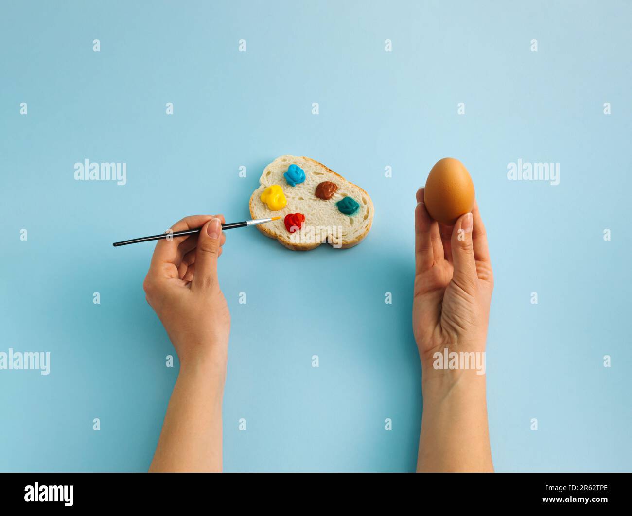 Un uovo bollito in mano e una fetta di pane con tavolozza colorata su sfondo blu pastello. La donna sta colorando le uova per la festa di Pasqua. Pasqua creativa c Foto Stock