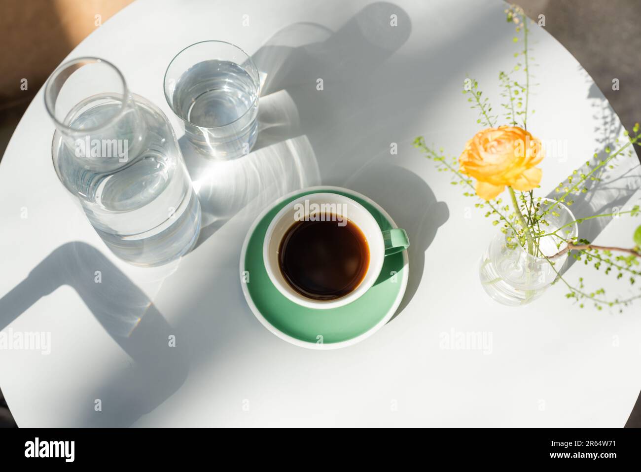 vista dall'alto di vetro e decanter con acqua pura, tazza con caffè nero, piattino, vaso con rosa gialla e piante verdi su tavola rotonda bianca al mattino Foto Stock