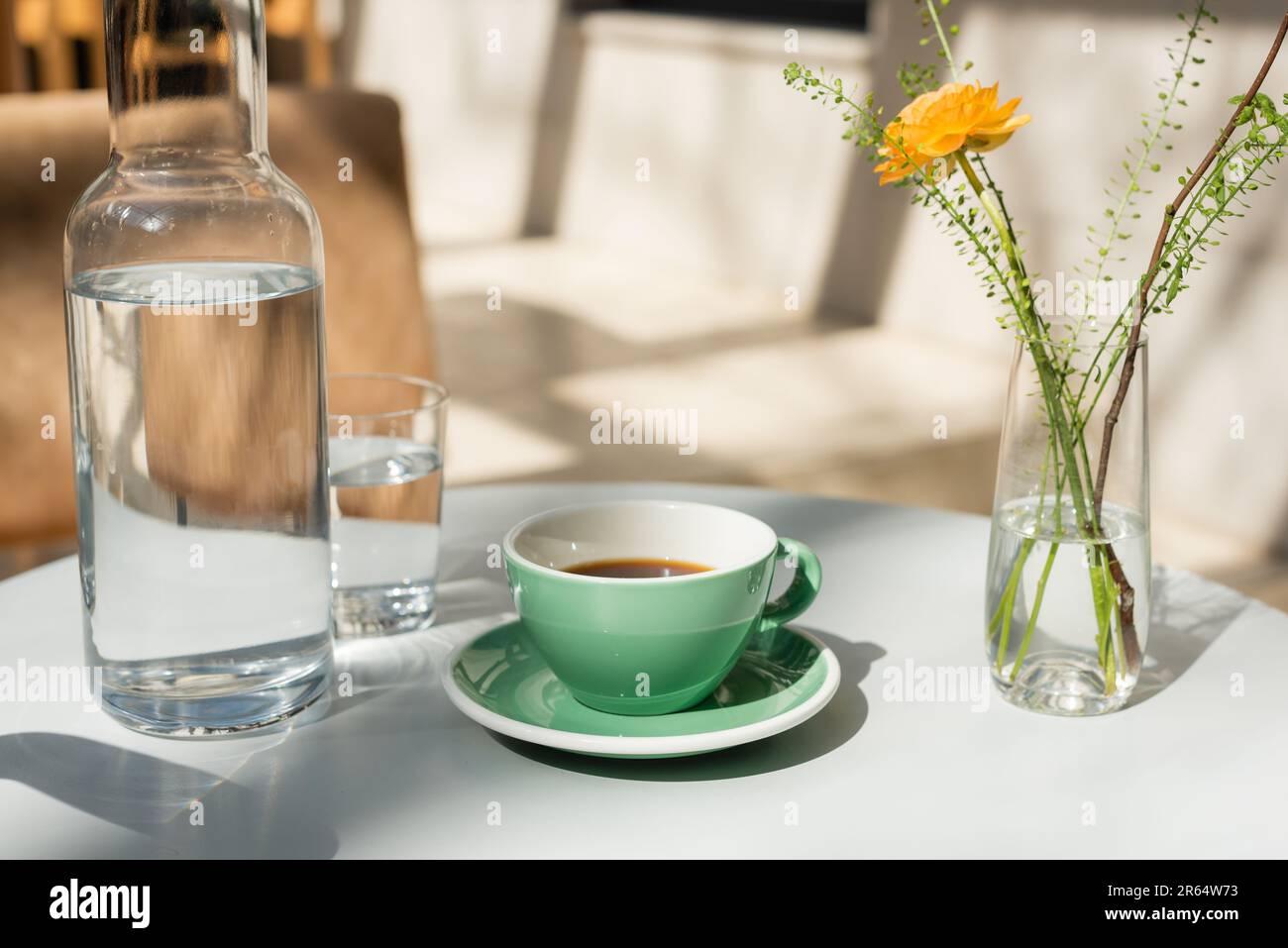 vaso con piante verdi e rosa gialle, bicchiere e decanter con acqua pura fresca, piattino, tazza con caffè nero su tavola rotonda bianca al mattino Foto Stock