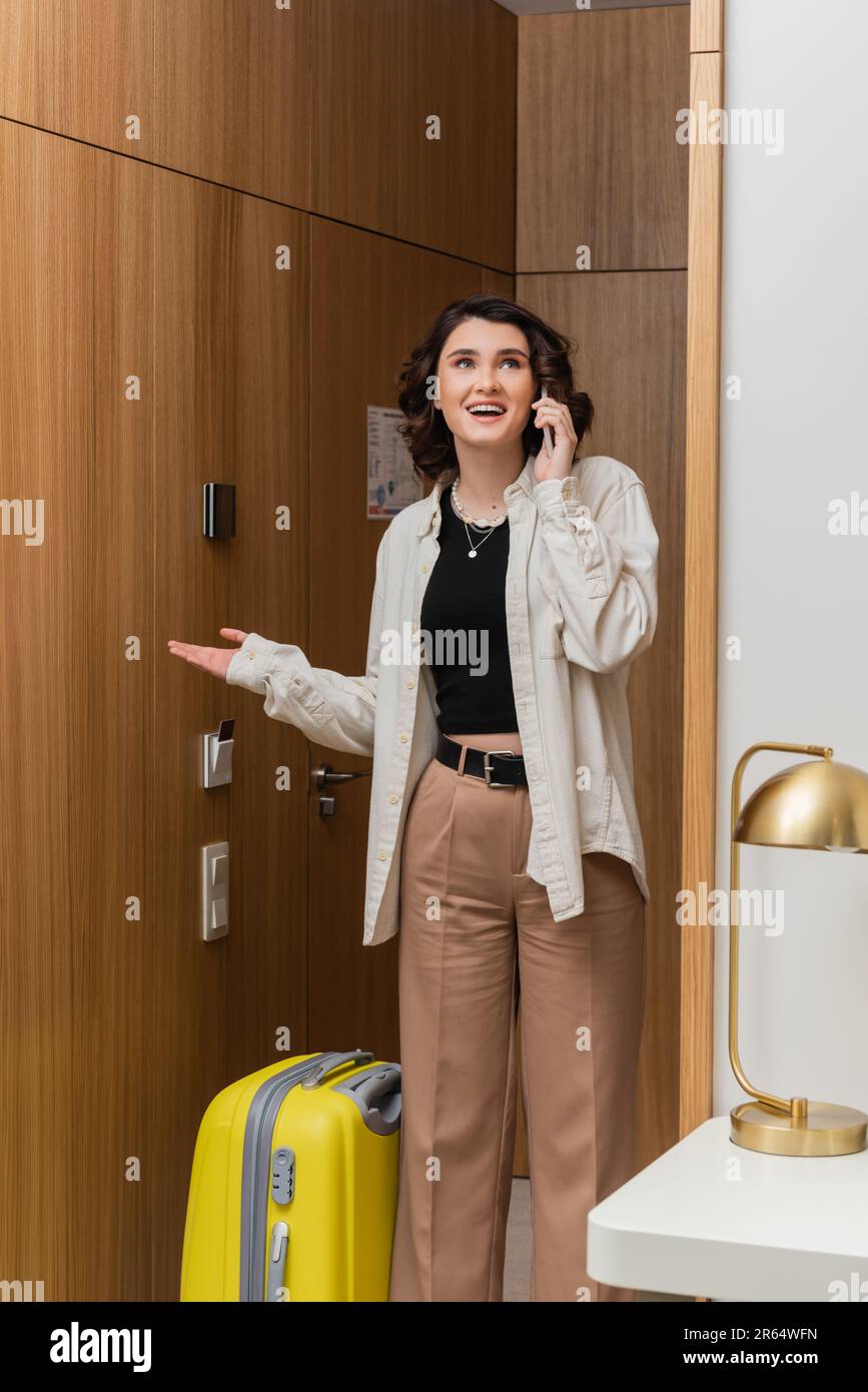 check-in, donna stupita con capelli brunette ondulati, in camicia bianca e pantaloni beige parlando sul telefono cellulare vicino alla porta d'ingresso con chiave elettronica, yel Foto Stock