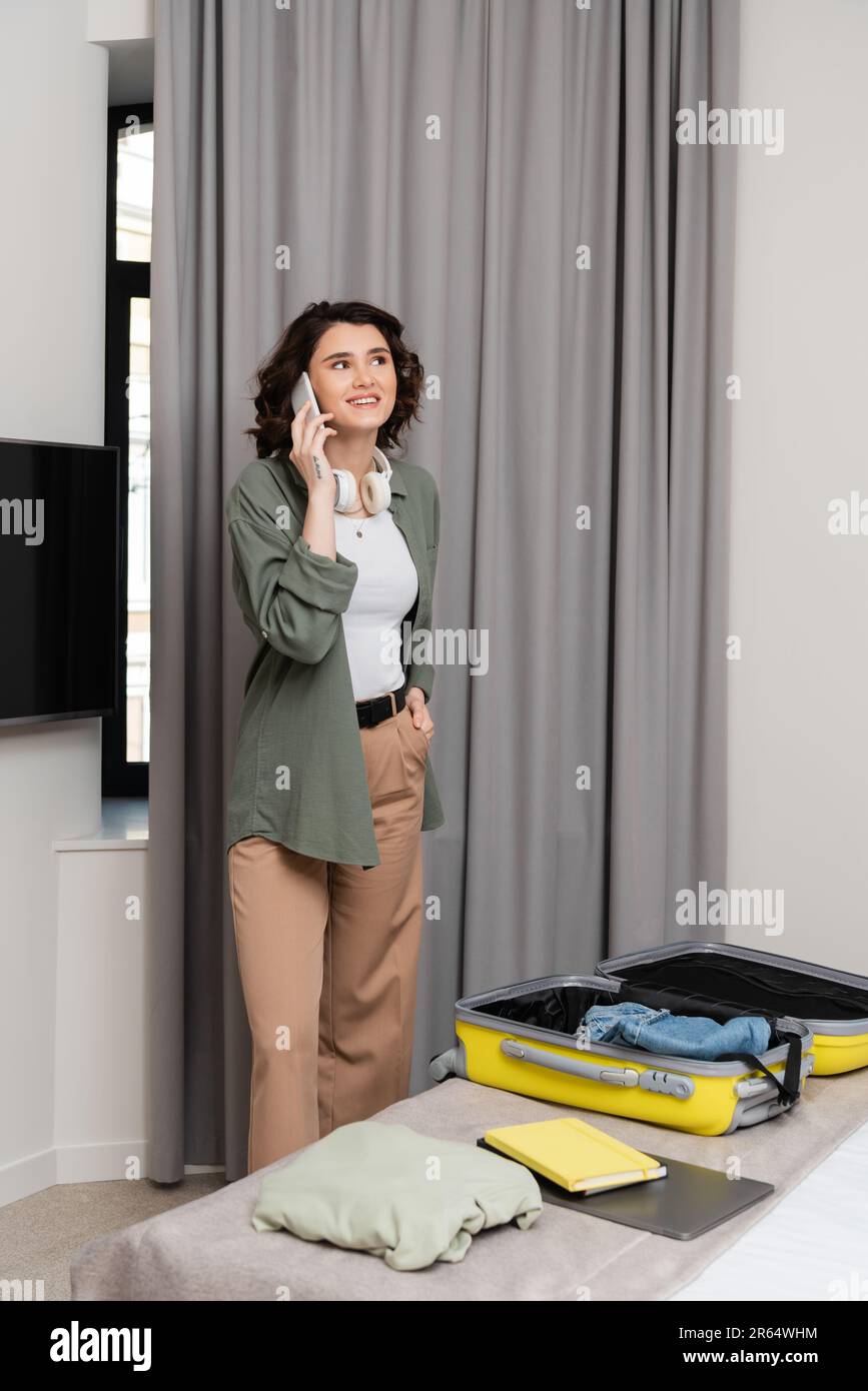 donna positiva e tatuata con cuffie senza fili che parlano sul telefono cellulare vicino a tende grigie, valigia con tutto il necessario per il viaggio, notebook e laptop Foto Stock
