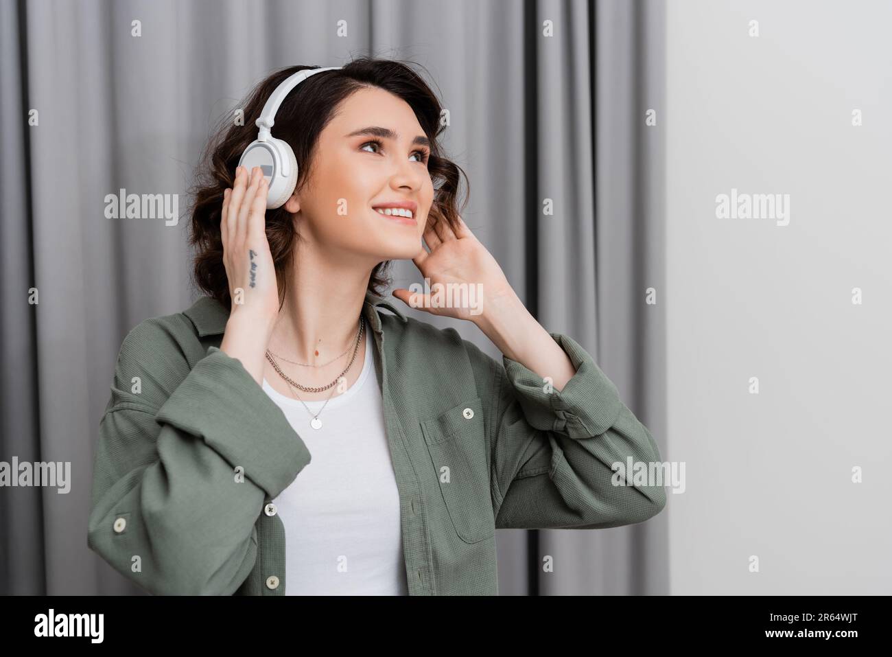 giovane donna entusiasta in abiti casual, con tatuaggio e capelli brunette ondulati guardando lontano mentre si ascolta la musica in cuffie wireless vicino grigio c Foto Stock