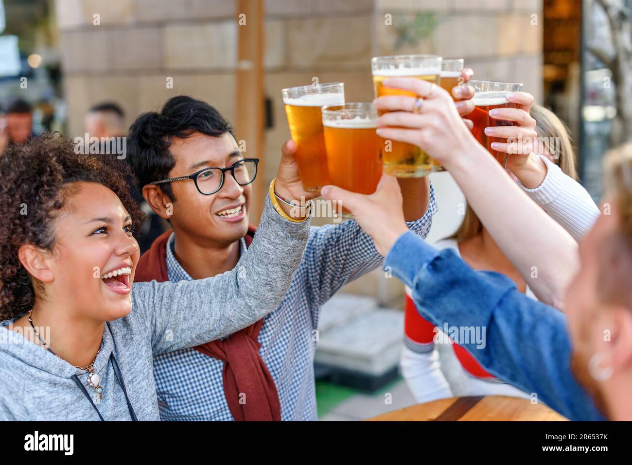 Gruppo di amici multirazziali positivi con il maschio raccolto in abiti casual sorridendo e guardando in su i bicchieri da birra e clinking mentre si è in piedi in legno ta Foto Stock