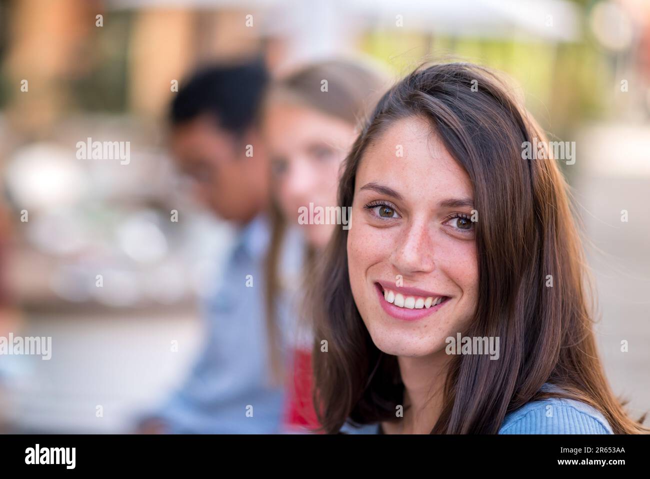 Giovane donna positiva in abiti casual e con lunghi capelli sorridenti e guardando la macchina fotografica mentre si trova in piedi in luce del giorno sulla strada della città contro Bloffed bac Foto Stock