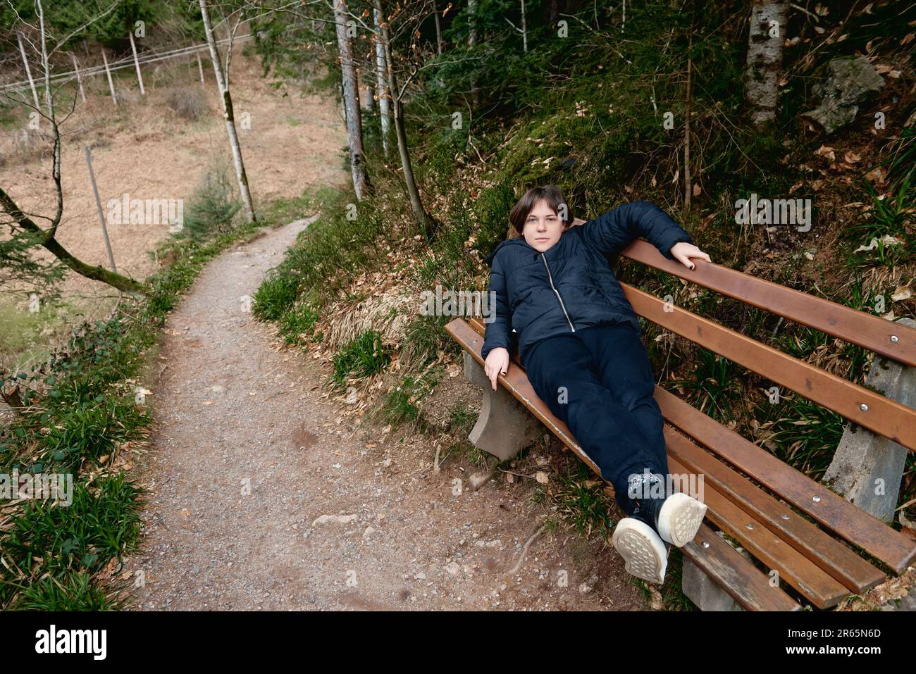 Bella giovane donna in abbigliamento sportivo sdraiata sulla panca di legno nel mezzo di un parco senza persone leggere un libro blu su uno sfondo di verde Foto Stock