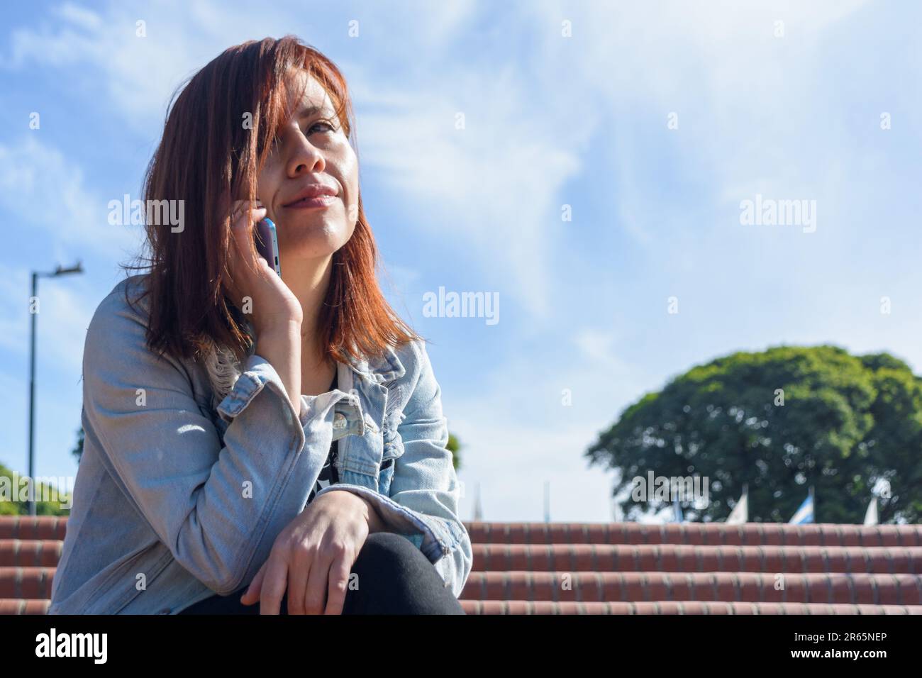 giovane colombiana latina in una telefonata seduta nel parco di buenos aires, con il cielo sullo sfondo e copia spazio, telefonia e technolo Foto Stock